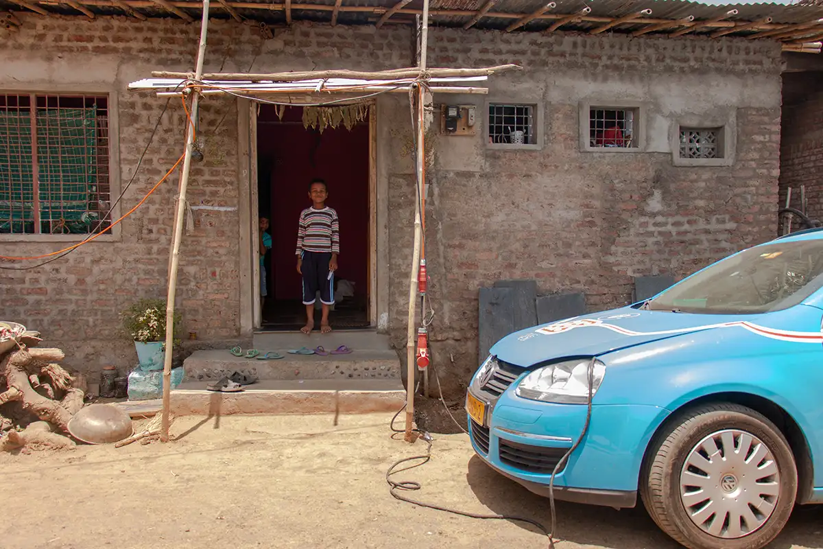 De Blue Bandit wordt opgeladen in een dorp in India. Een lokale bewoner kijkt toe vanaf zijn voordeur.